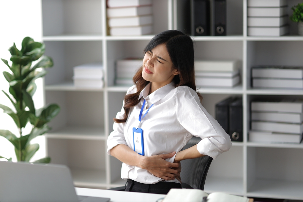 Woman experiencing back pain while sitting at her desk in an office, highlighting the impact of back pain at work and the need for physiotherapy.